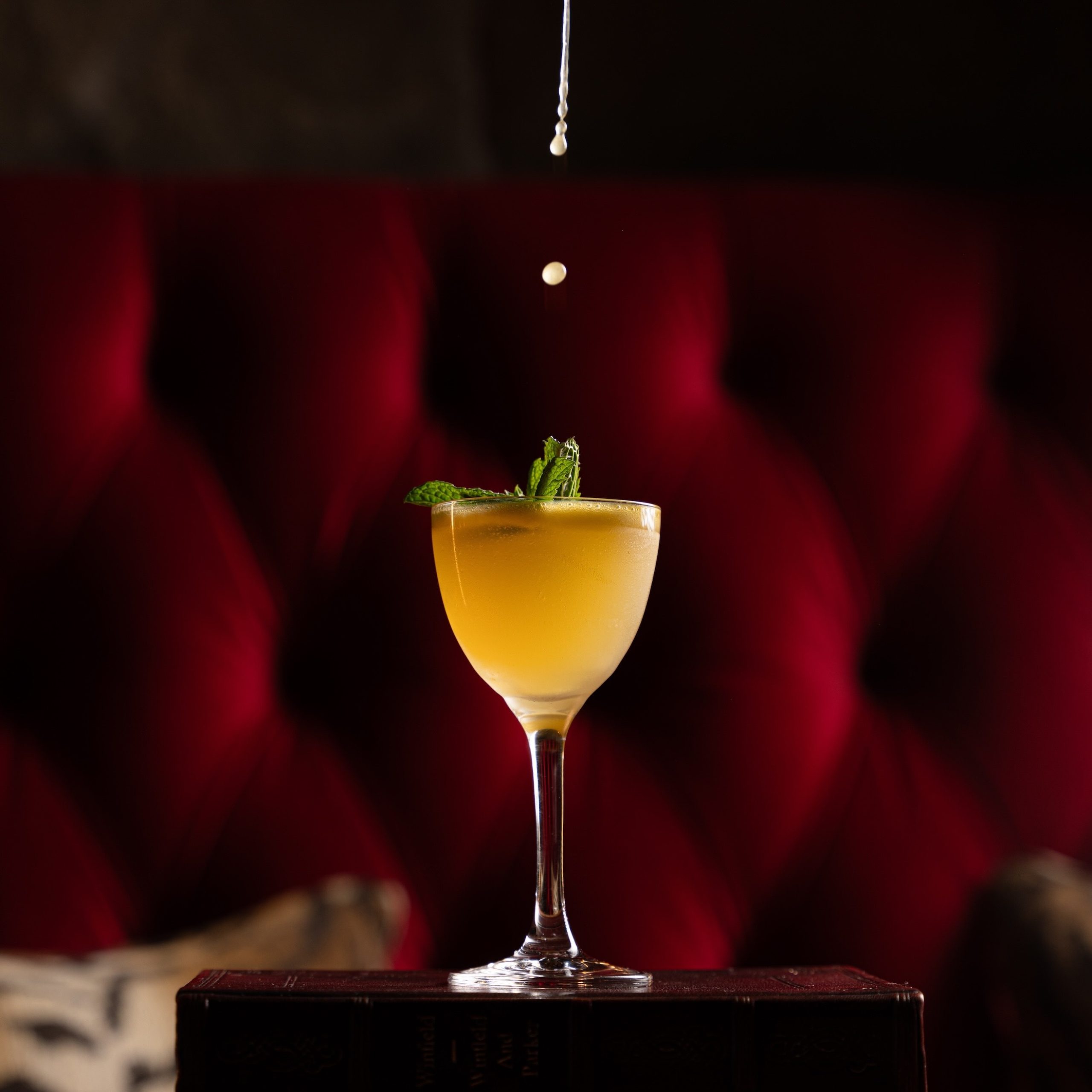 A beautifully presented cocktail garnished with mint, resting on stacked books against a rich red background.