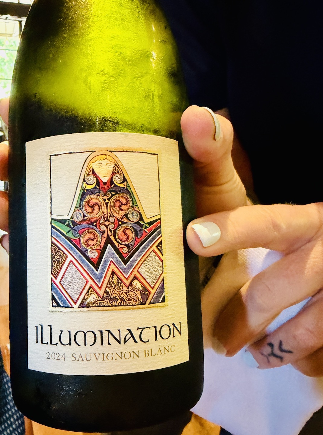 A hand holds a bottle of Illumination 2024 Sauvignon Blanc featuring intricate label artwork.
