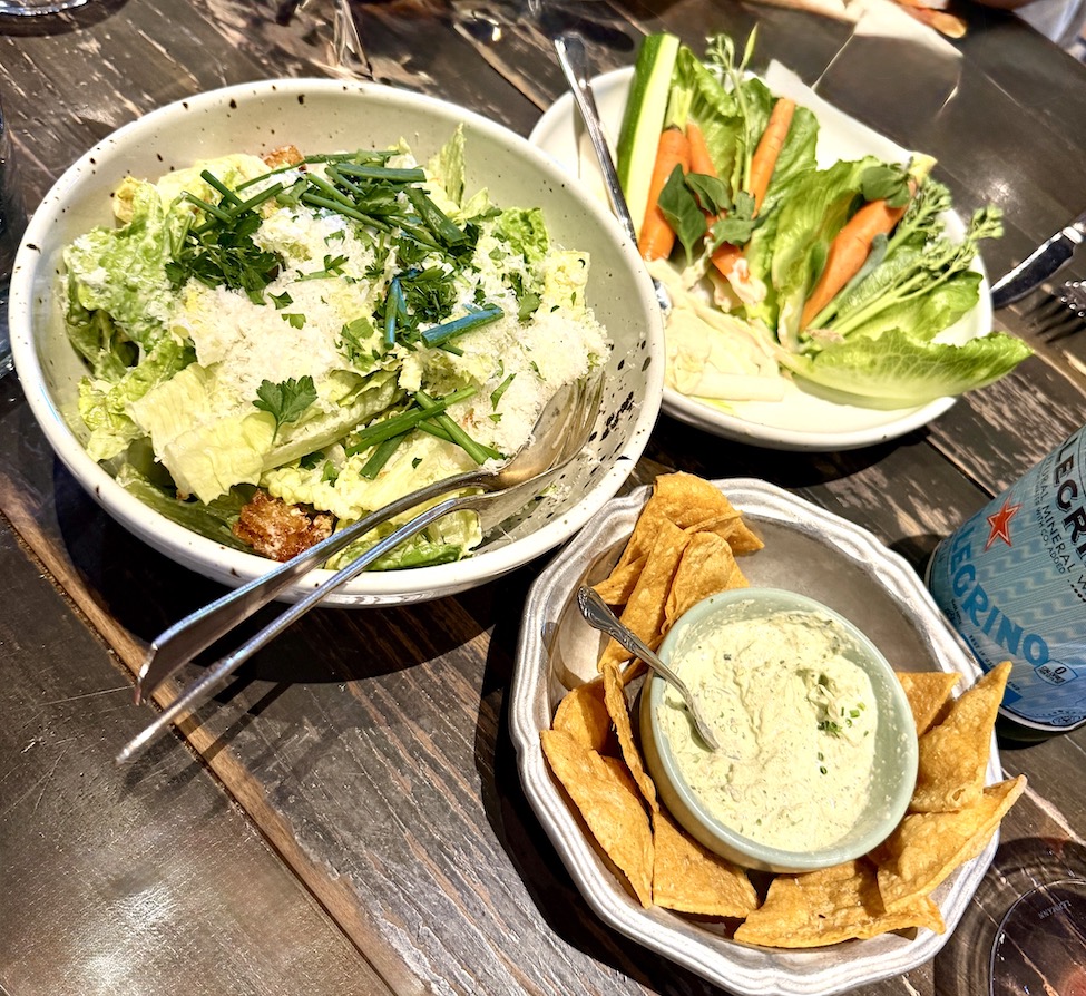 A bowl of Caesar salad topped with herbs and cheese, accompanied by a platter of fresh vegetables and tortilla chips with dip.