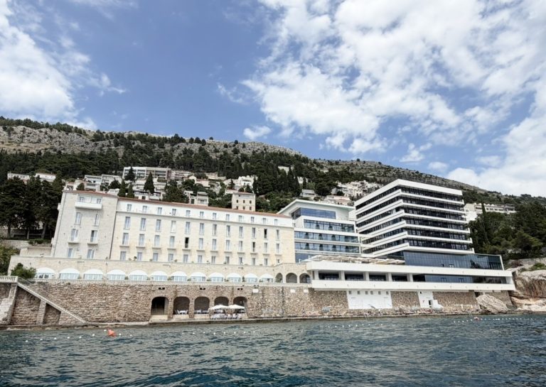 Hotel Excelsior Dubrovnik overlooking the Adriatic Sea, with a backdrop of lush hills and a partly cloudy sky.