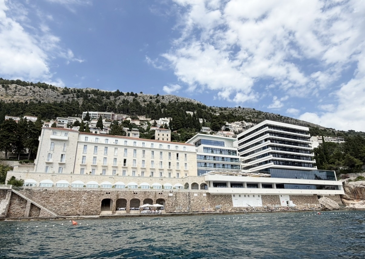 Hotel Excelsior Dubrovnik overlooking the Adriatic Sea, with a backdrop of lush hills and a partly cloudy sky.