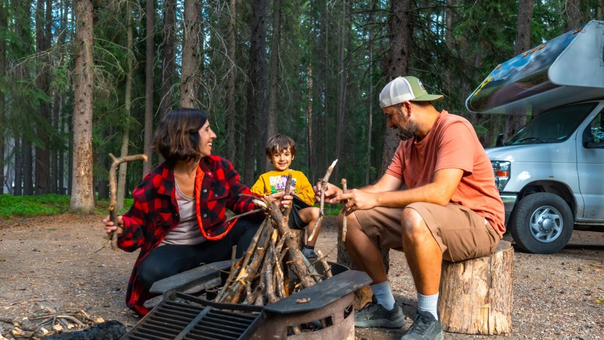 Family gathered around a campfire in a forest, enjoying time together near a campervan.
