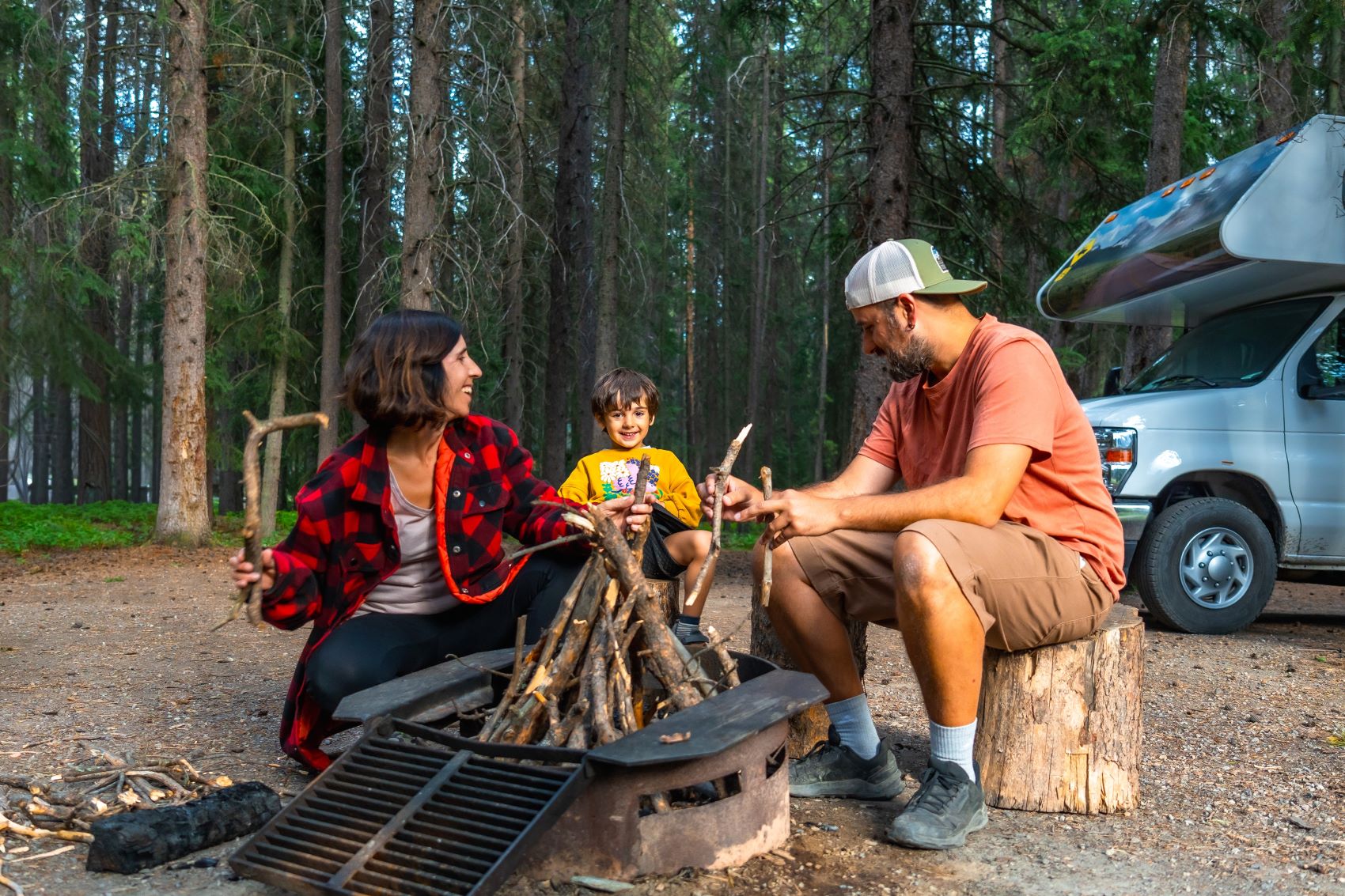 Family gathered around a campfire in a forest, enjoying time together near a campervan.