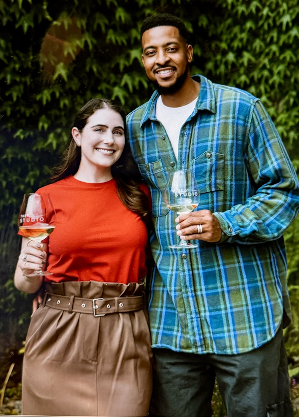 Elise and CJ McCollum smile while holding glasses of wine, set against a lush green background.