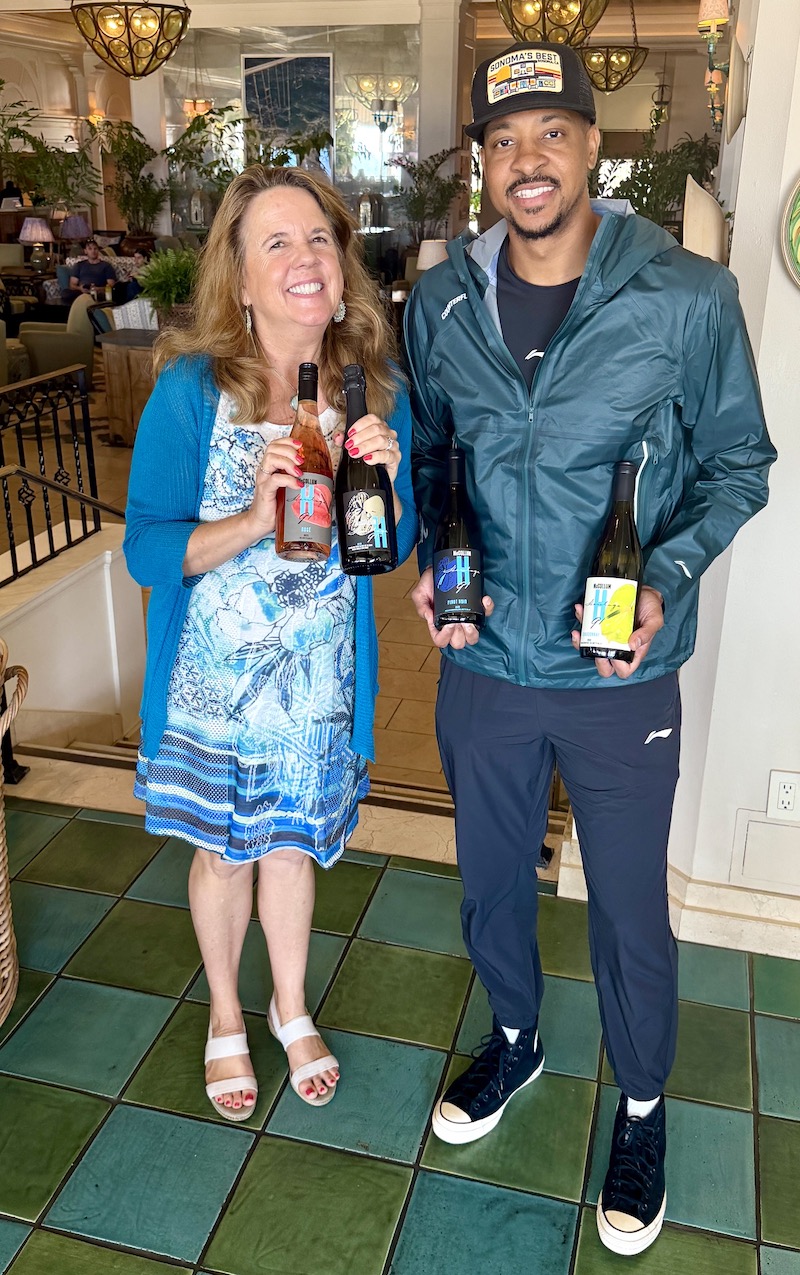 Jill Weinlein and CJ McCollum pose together, holding various bottles of wine in a stylish indoor setting.