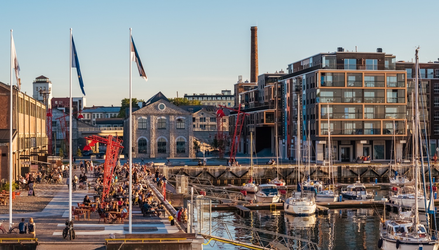 Crowds enjoy the waterfront at Noblessner, surrounded by modern buildings, boats, and flags against a clear sky.