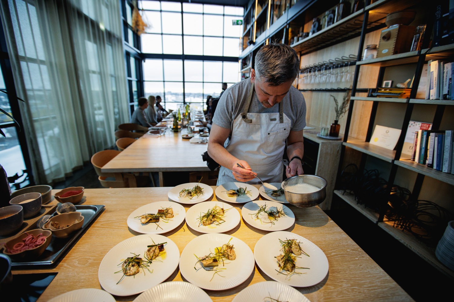 A chef plating dishes with seafood and garnishes at a modern dining table with a bright window view.