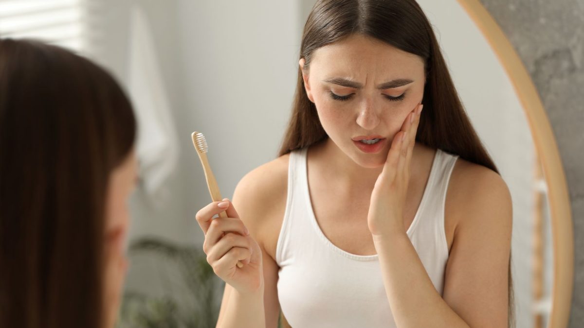 Young woman holding a toothbrush, looking concerned about tooth pain while examining herself in the mirror.