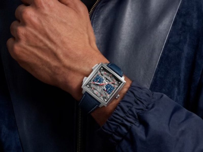 A stylish hand wearing a modern luxury watch with a square face and blue strap against a dark jacket backdrop.