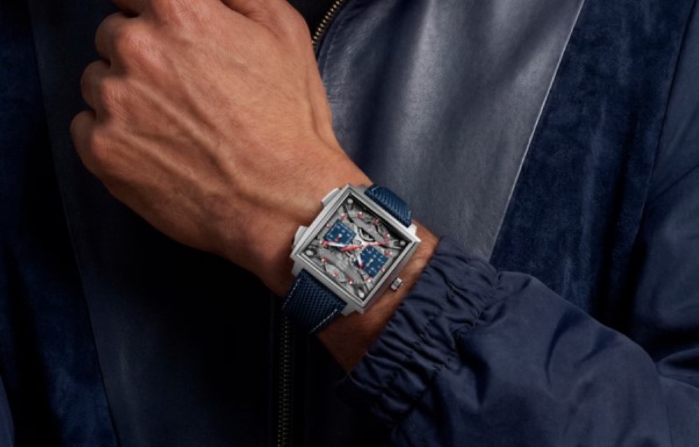 A stylish hand wearing a modern luxury watch with a square face and blue strap against a dark jacket backdrop.