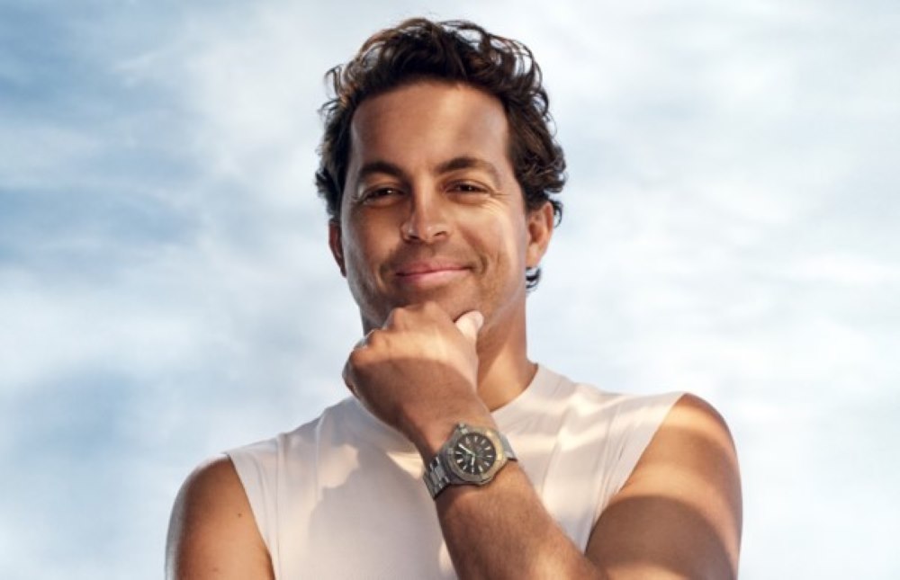 Man in a sleeveless shirt poses confidently while showcasing a modern luxury watch against a soft sky backdrop.