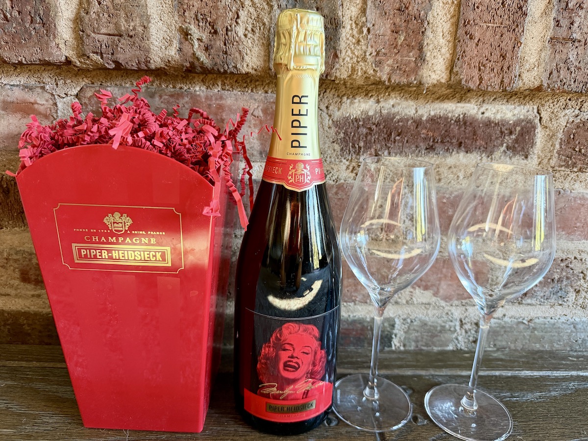 Piper-Heidsieck champagne bottle with Marilyn Monroe label, surrounded by decorative red paper and two wine glasses.