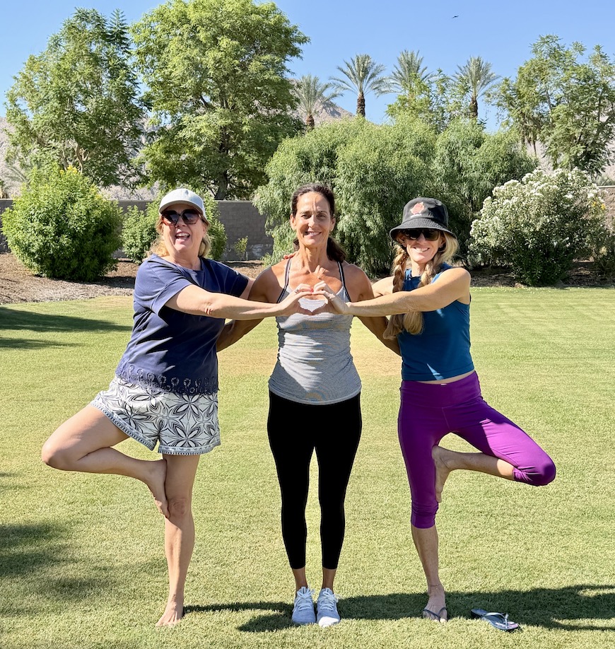 Three women doing yoga poses while forming a heart shape with their hands on a sunny lawn at a resort.
