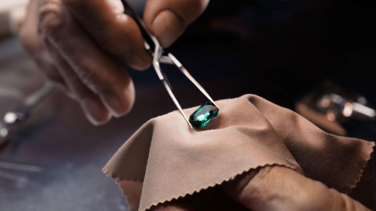A jeweler using tweezers to hold a green gemstone over a soft cloth during a jewelry repair process.