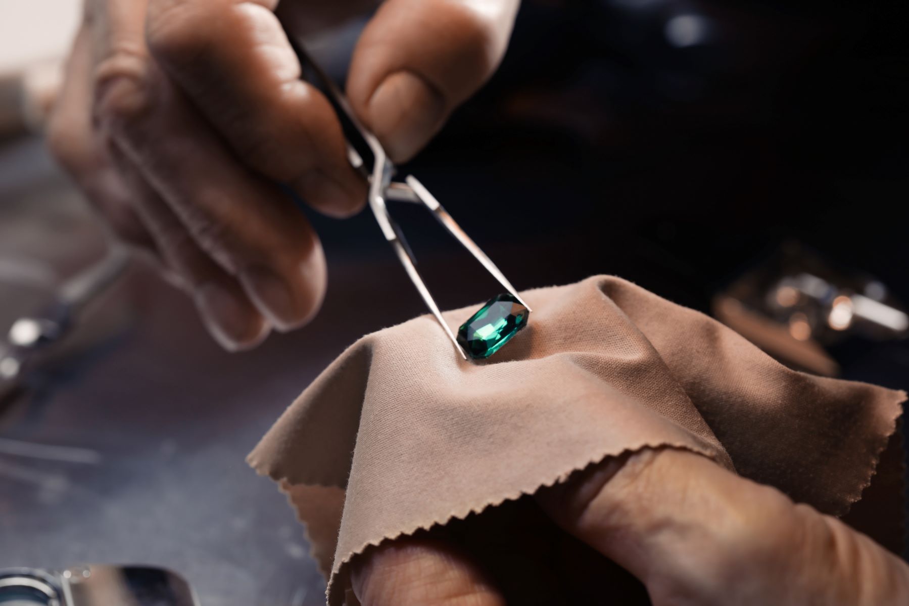 A jeweler using tweezers to hold a green gemstone over a soft cloth during a jewelry repair process.