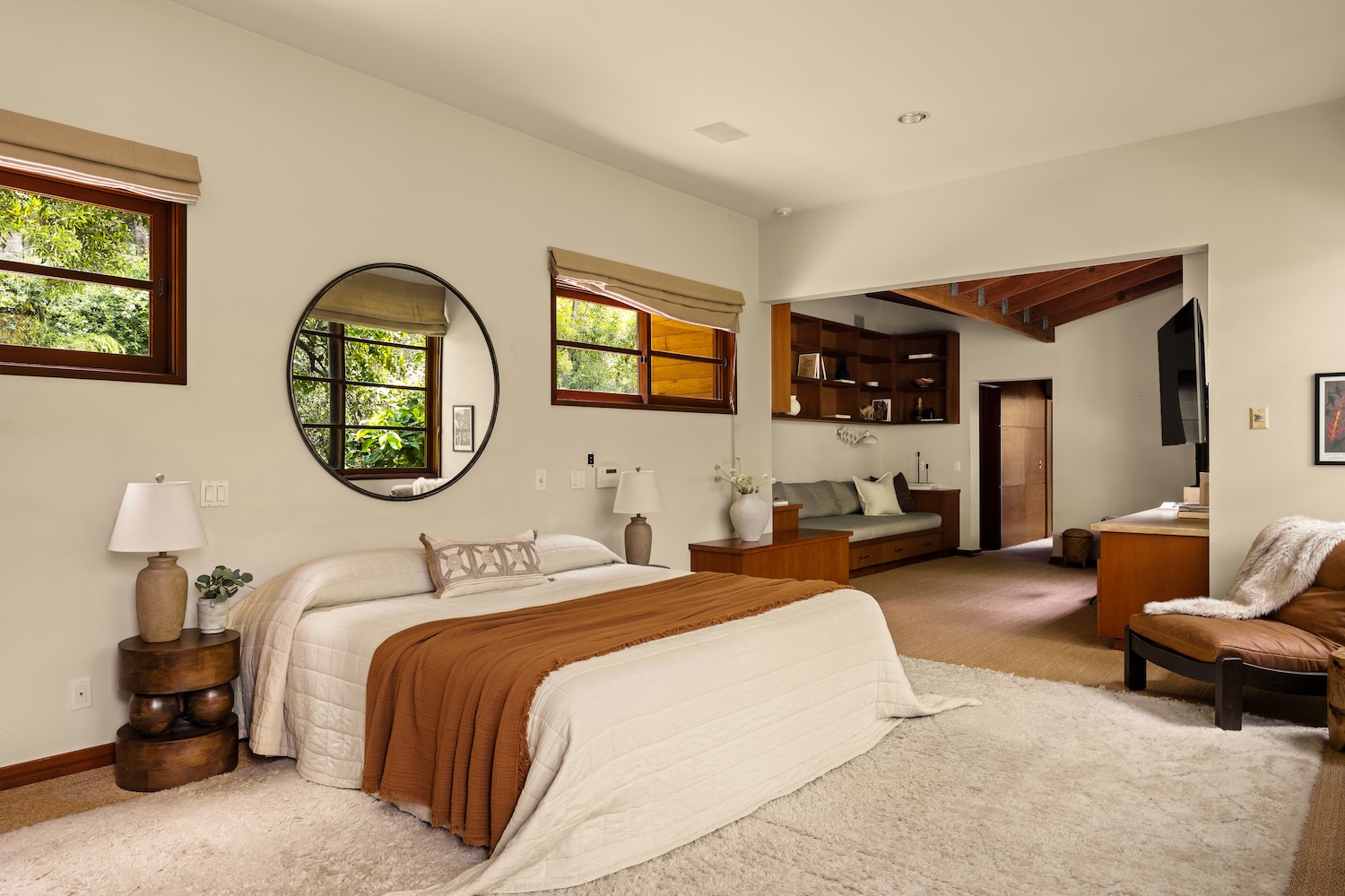 Cozy bedroom featuring a bed with an earthy blanket, circular mirror, natural light from windows, and stylish wooden furniture.