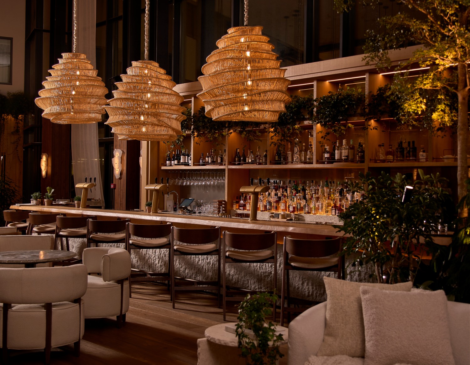 Elegant bar area featuring unique hanging lights, a variety of spirits on display, and lush greenery in a cozy atmosphere.