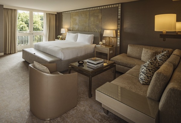 Luxurious hotel room featuring a king bed, modern decor, a sitting area with plush furniture, and natural light from large windows.