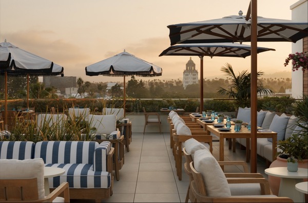 Rooftop dining area at L’Ermitage Beverly Hills with striped furniture, palm plants, and a view of a distant tower at sunset.
