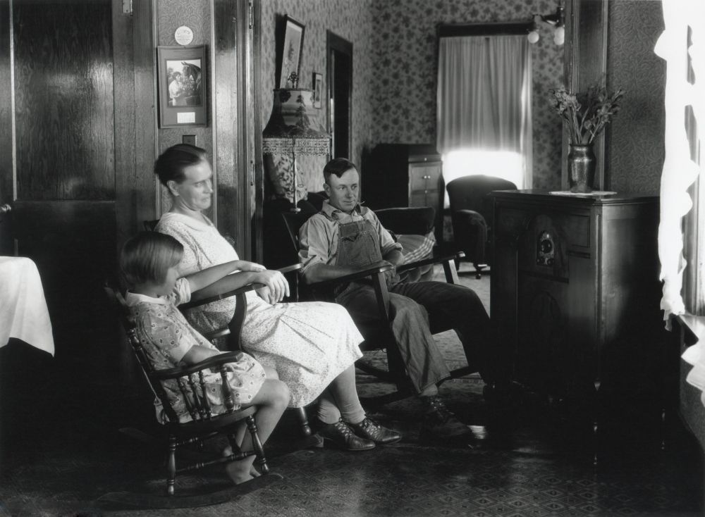 A woman, girl, and man sit in a warmly decorated room, engaged in family time around an old radio.