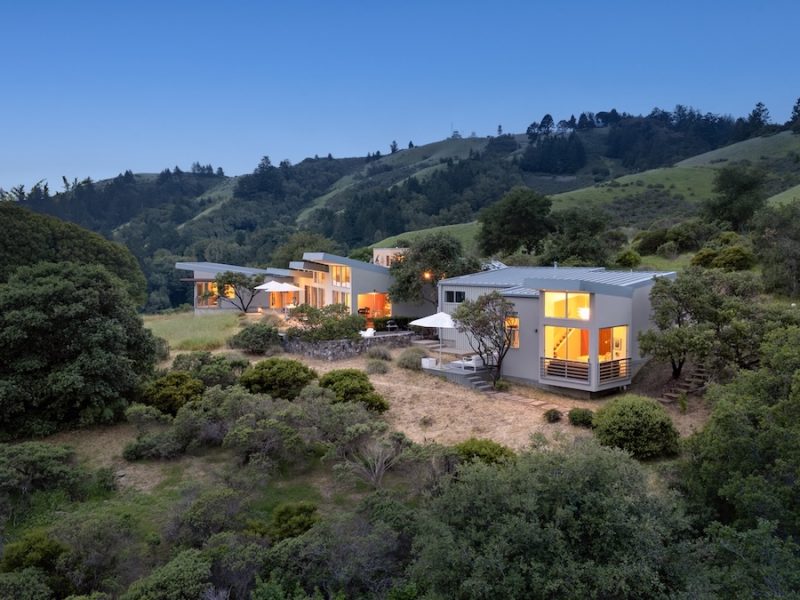 Modern retreat houses nestled in the hills of West Marin, surrounded by lush greenery and a clear evening sky.