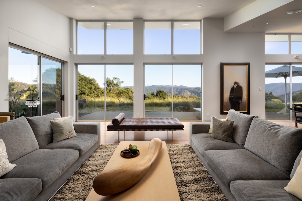 Modern living room with large windows showcasing views of hills and greenery, featuring comfortable sofas and a wooden coffee table.