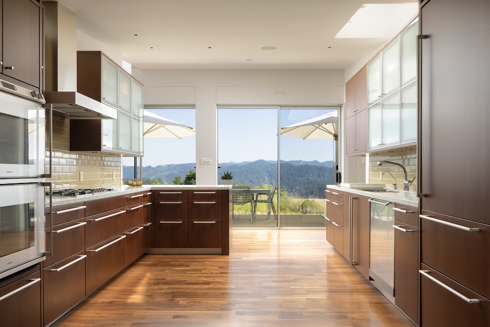 Modern kitchen with wood cabinetry and a view of hills, featuring large windows and a patio with outdoor seating.