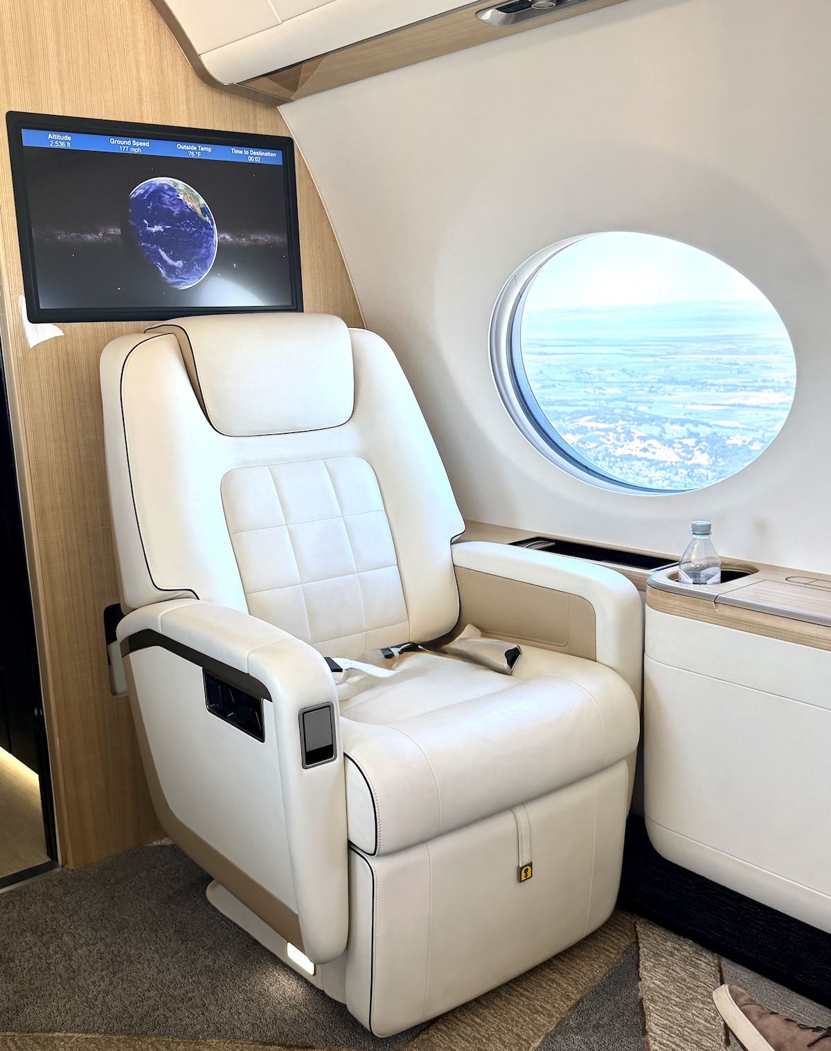 Luxurious white leather seat in a Gulfstream G500 jet with a monitor displaying flight information and a circular window view.
