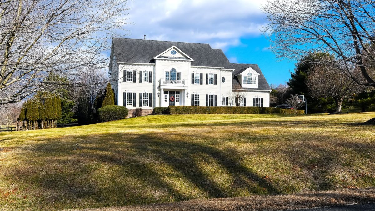 Elegant white colonial-style house with manicured lawn in a wealthy neighborhood under a blue sky with scattered clouds.
