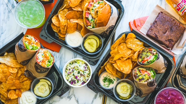 A variety of sandwiches, potato chips, salads, pickles, and desserts arranged in takeout boxes, ideal for summer gatherings.