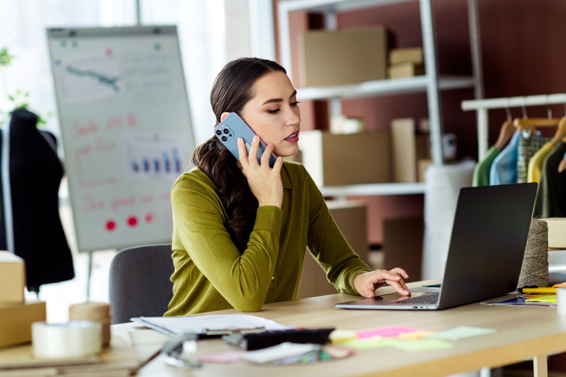 A woman in a green sweater talks on the phone while working on a laptop in a stylish boutique environment.