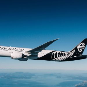 Air New Zealand widebody aircraft soaring above a picturesque landscape with mountains and water below.
