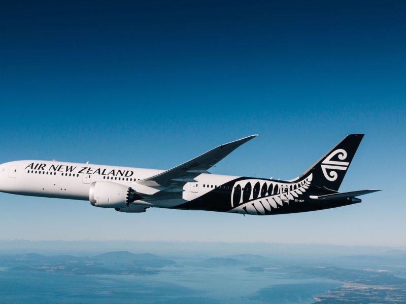 Air New Zealand widebody aircraft soaring above a picturesque landscape with mountains and water below.