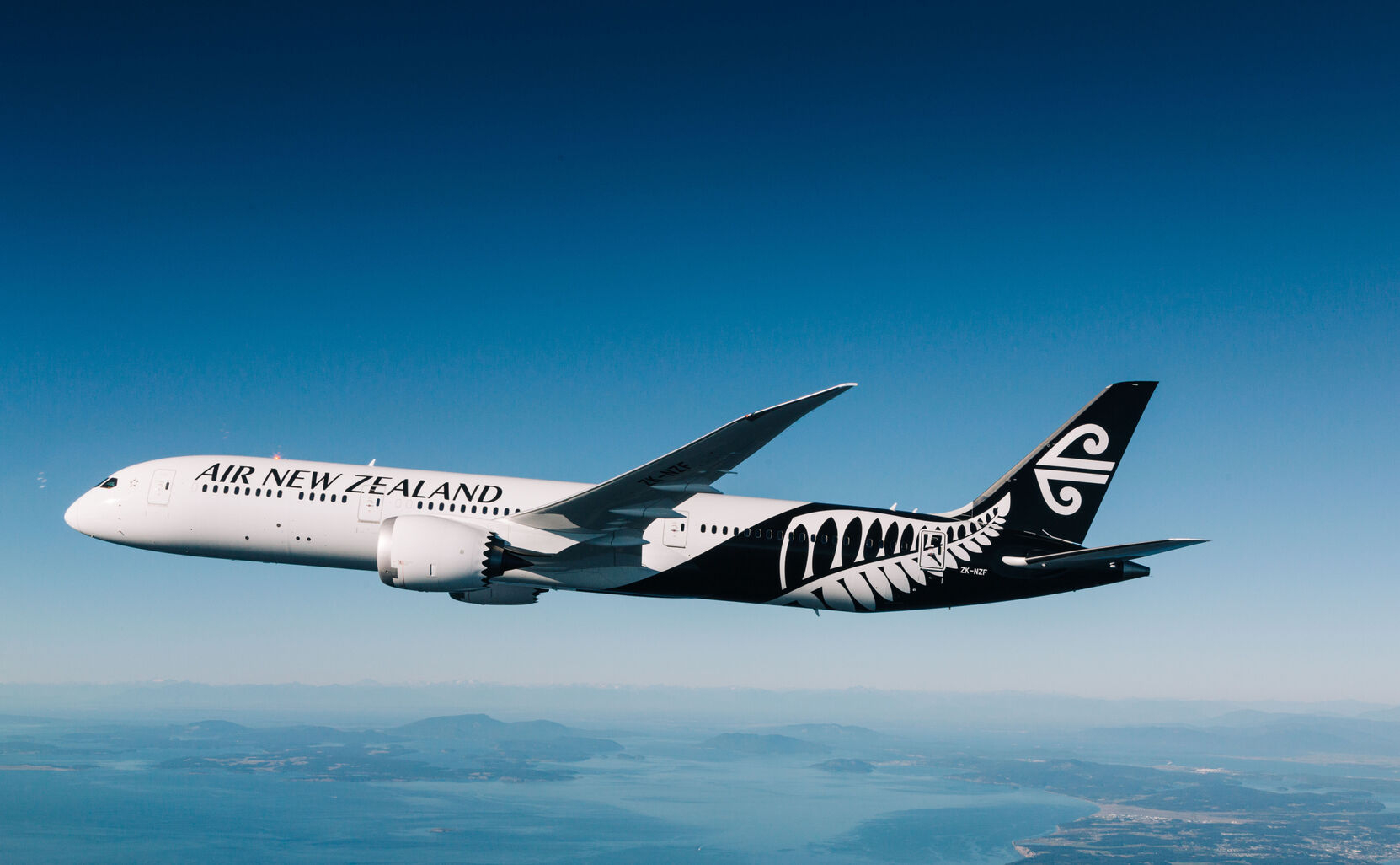 Air New Zealand widebody aircraft soaring above a picturesque landscape with mountains and water below.