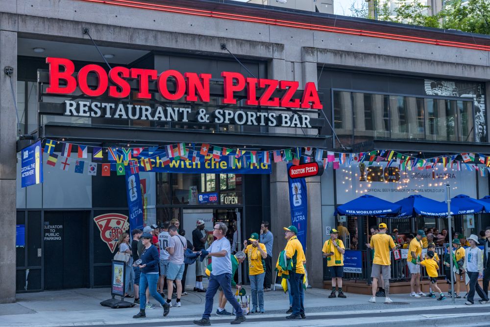 Crowd outside Boston Pizza restaurant, decorated with flags, celebrating FIFA World Cup 2026 hospitality events and experiences.