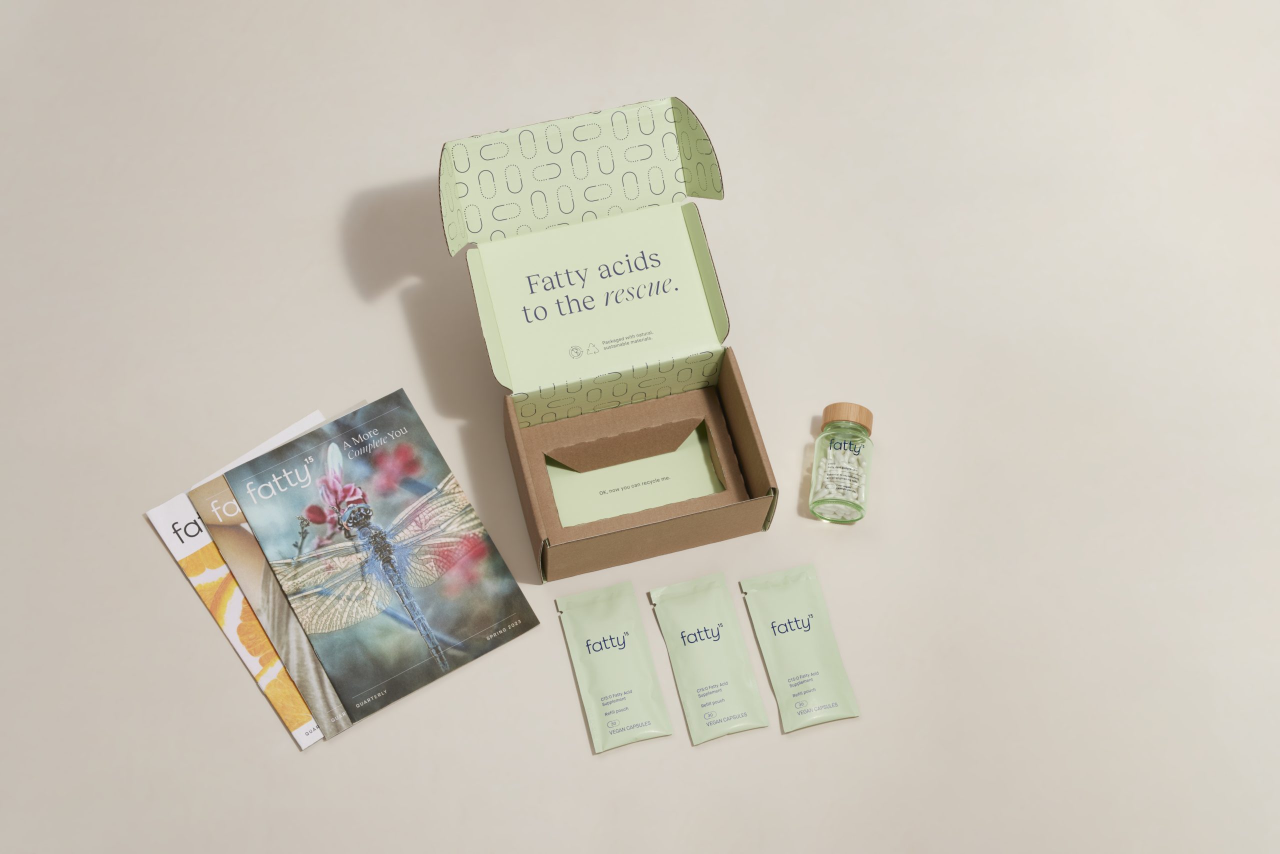 A subscription box open to reveal C15 fatty acid supplements, brochures, and a glass bottle alongside packaged samples.