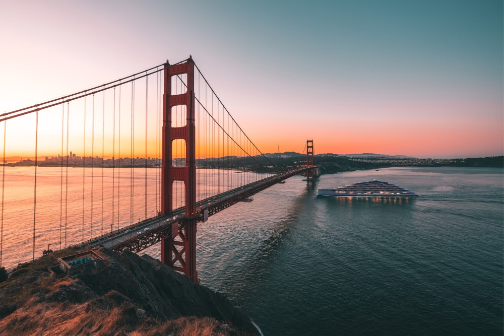 Golden Gate Bridge spans the bay at sunset, with a boat navigating the calm waters and San Francisco in the background.