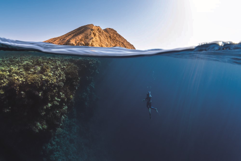 A diver exploring underwater coral while a mountainous island rises above the water surface.