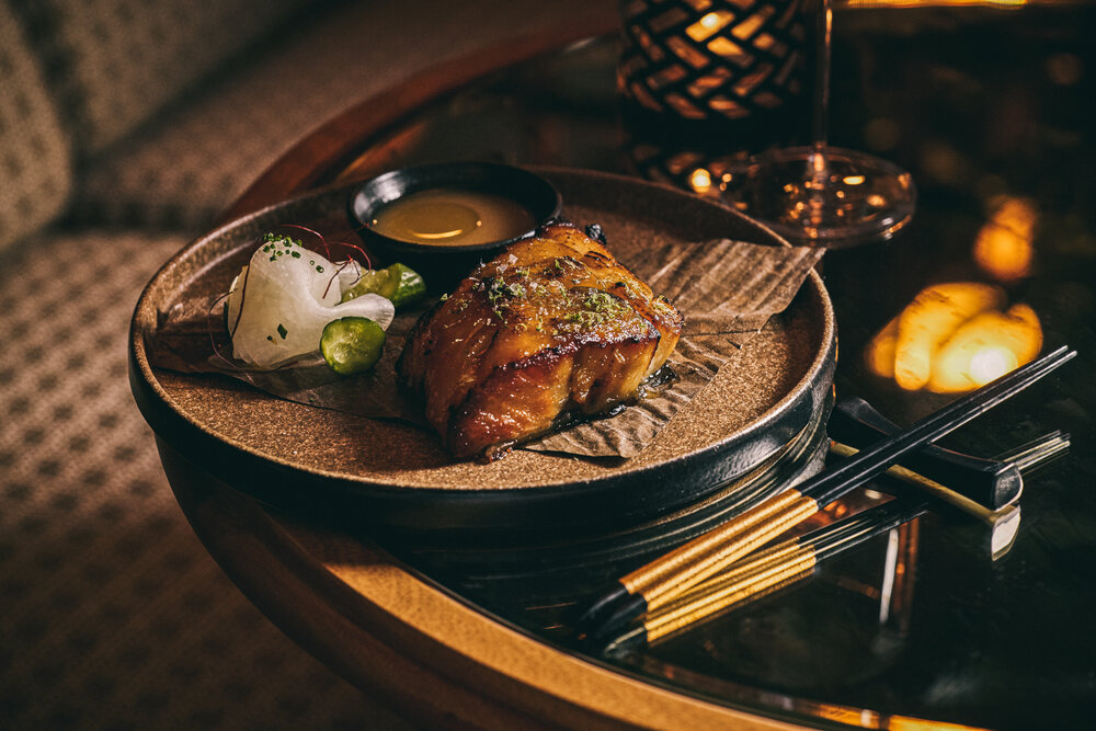 Glazed black cod fillet served on a decorative plate with garnishes and dipping sauce, accompanied by chopsticks.