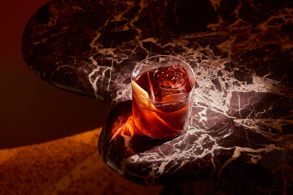 A cocktail glass with ice and a citrus twist sits on a dark marble table, illuminated by warm light.