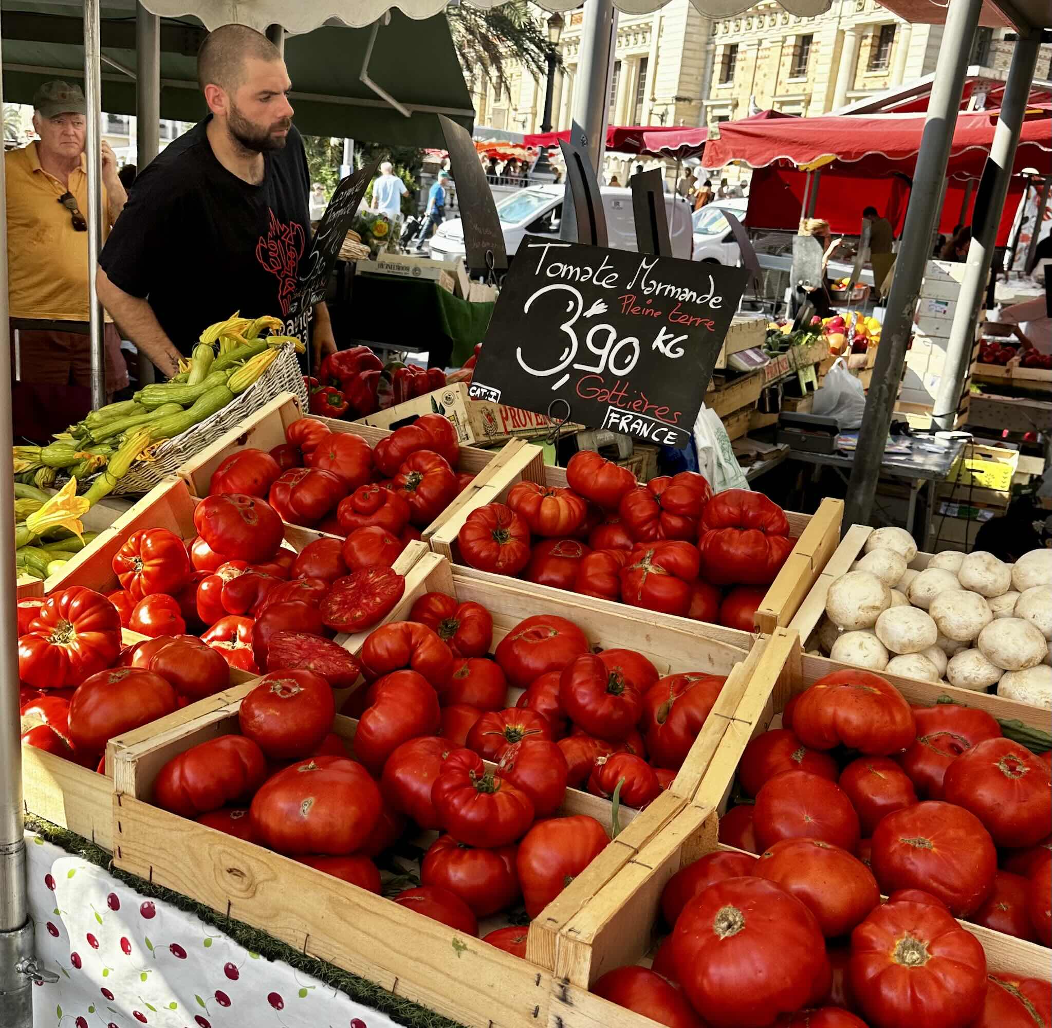 A vendor displays vibrant red tomatoes at a market stall with a sign listing prices and origins in Nice.