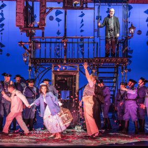 A lively scene from Il Barbiere di Siviglia, featuring performers in colorful costumes amid a burst of petals.