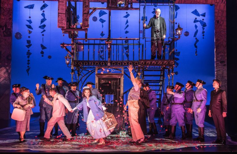 A lively scene from Il Barbiere di Siviglia, featuring performers in colorful costumes amid a burst of petals.