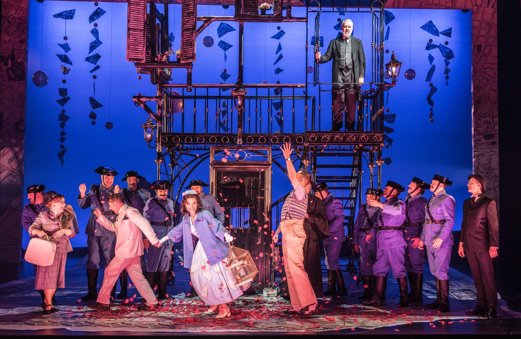 A lively scene from Il Barbiere di Siviglia, featuring performers in colorful costumes amid a burst of petals.