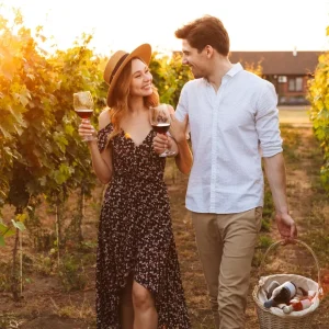Couple enjoying wine in a vineyard during sunset, holding glasses and walking together, with a basket of wine bottles nearby.