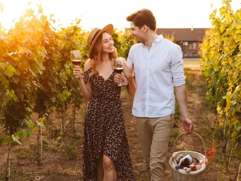 Couple enjoying wine in a vineyard during sunset, holding glasses and walking together, with a basket of wine bottles nearby.