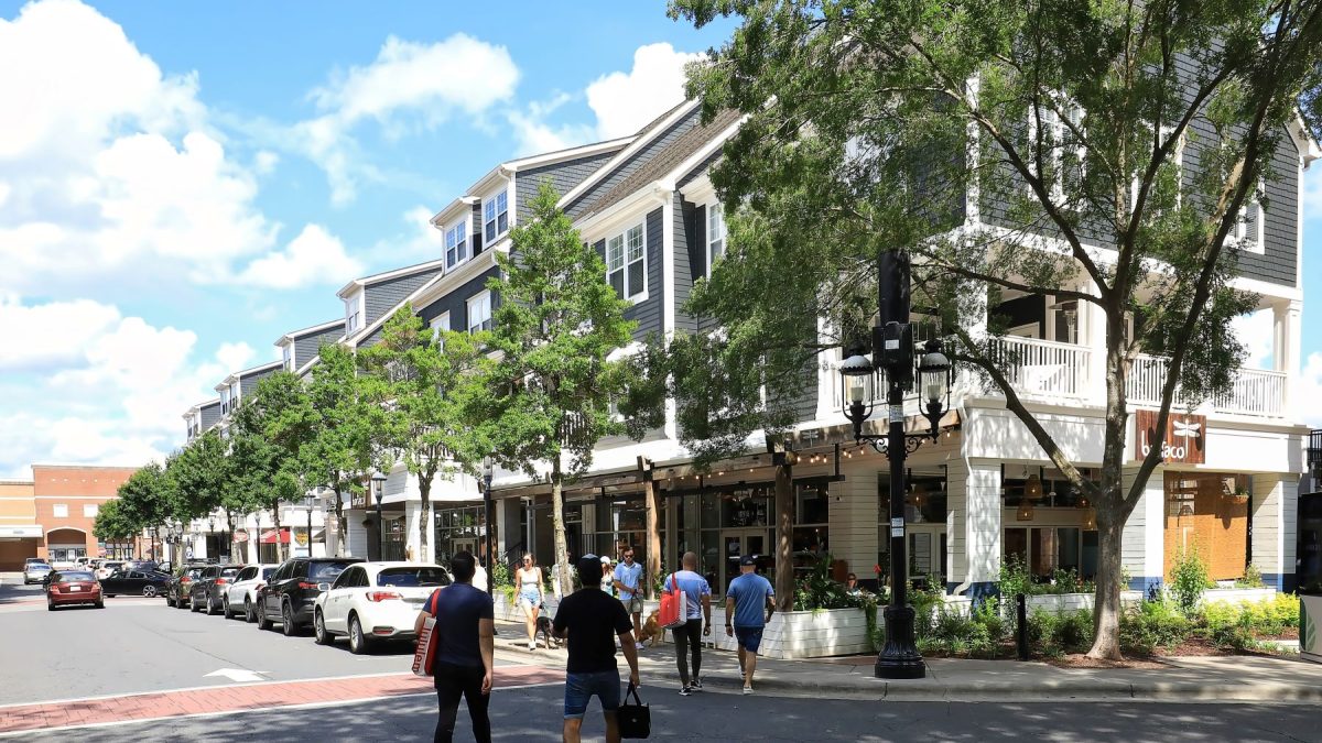 Crowds walk along a tree-lined street in Huntersville, featuring shops and sunny skies.