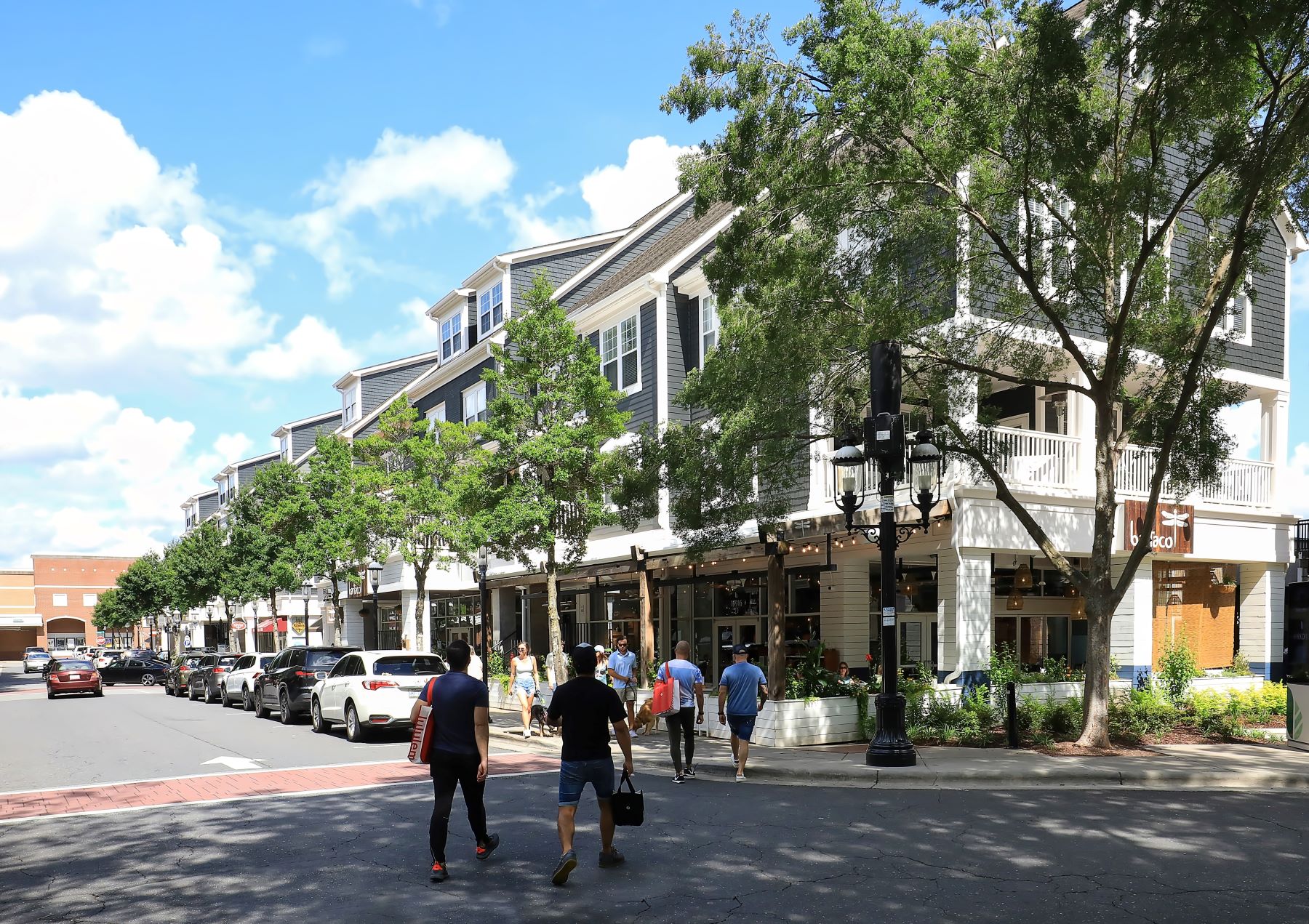 Crowds walk along a tree-lined street in Huntersville, featuring shops and sunny skies.
