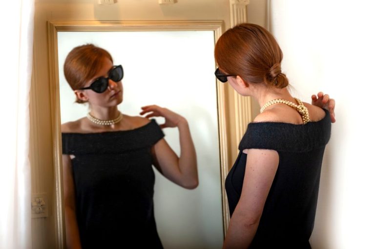 Woman in black evening gown adjusting her hair and looking at her reflection in a mirror, wearing sunglasses and a pearl necklace.