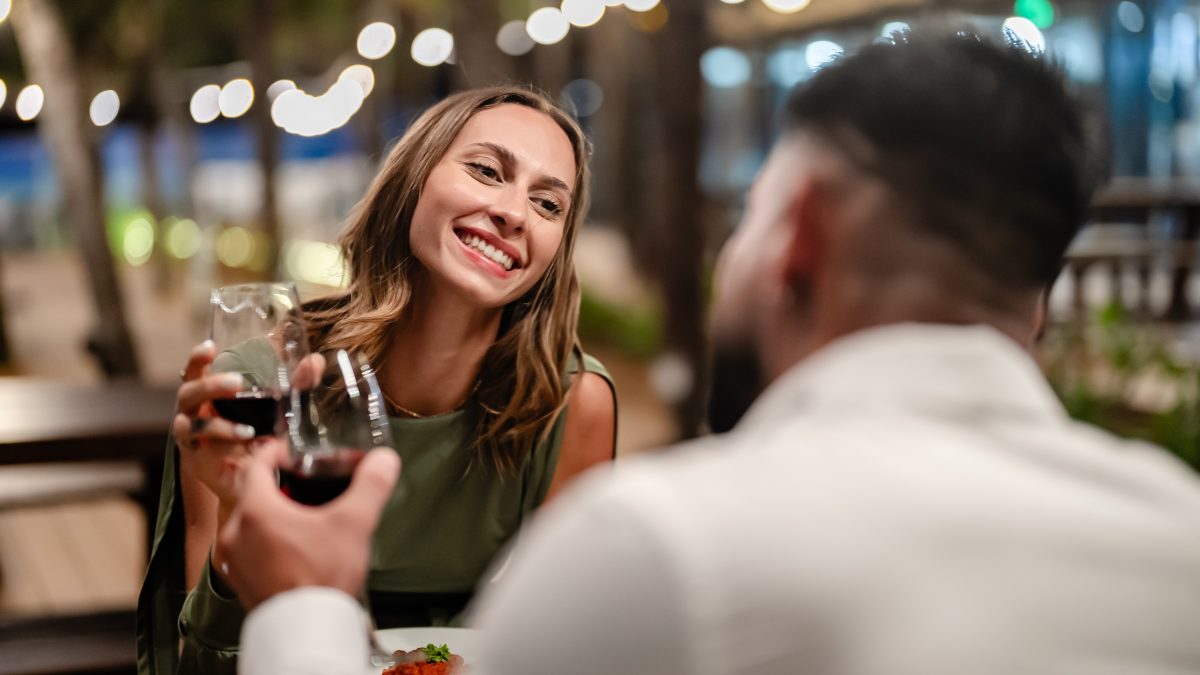 A couple enjoying a dinner date with glasses of red wine, smiling at each other in a cozy outdoor setting.